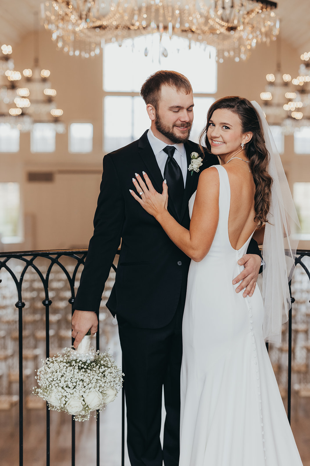 Nichole and Gavin on a balcony beneath lit chandeliers, her veil falling over a sleek crepe gown and a bouquet of baby's breath in his hand